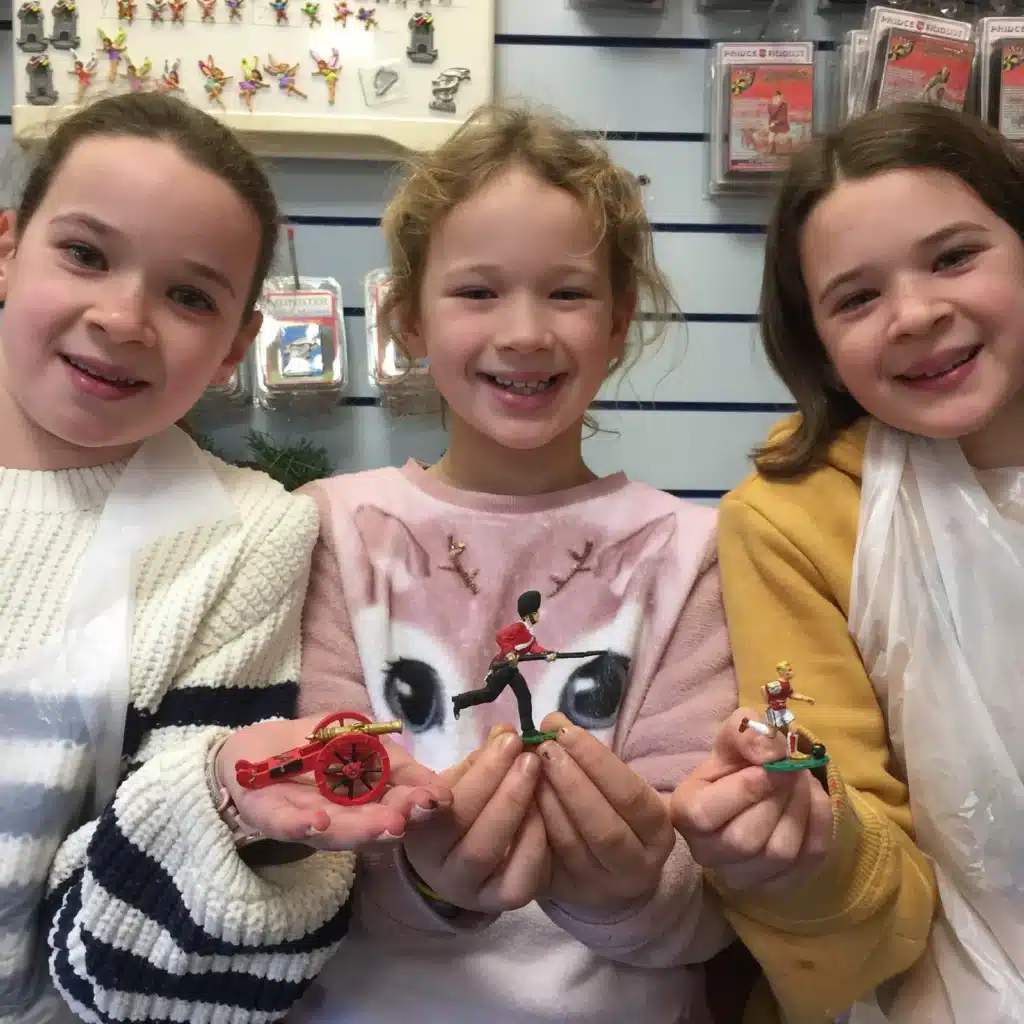 Three smiling children hold up collectible soldier figurines in a shop. They're wearing casual clothing and sitting in front of a display filled with various small items. The children appear to be excited about their collections.