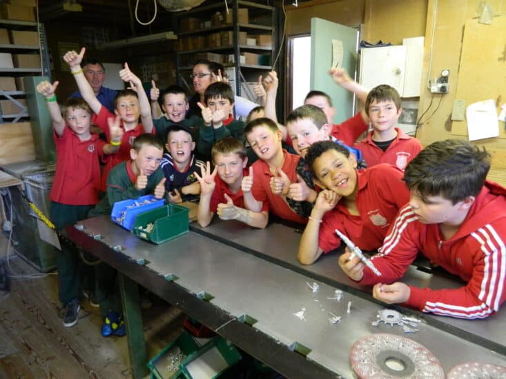 A group of children in colorful uniforms are gathered around a workbench in a workshop. They are smiling, giving thumbs-up, and appear excited. Some are holding small metal objects. An adult in the background watches over them. Various tools are visible on the bench.