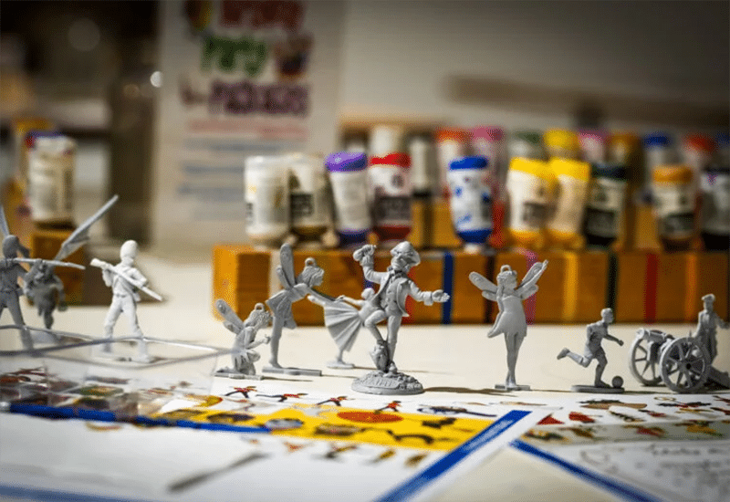 A table is filled with unpainted miniature figurines of various characters, including fairies and soldiers. Paintbrushes, colorful paint tubes arranged in rows, and sheets of example designs are visible in the background, suggesting a painting activity.