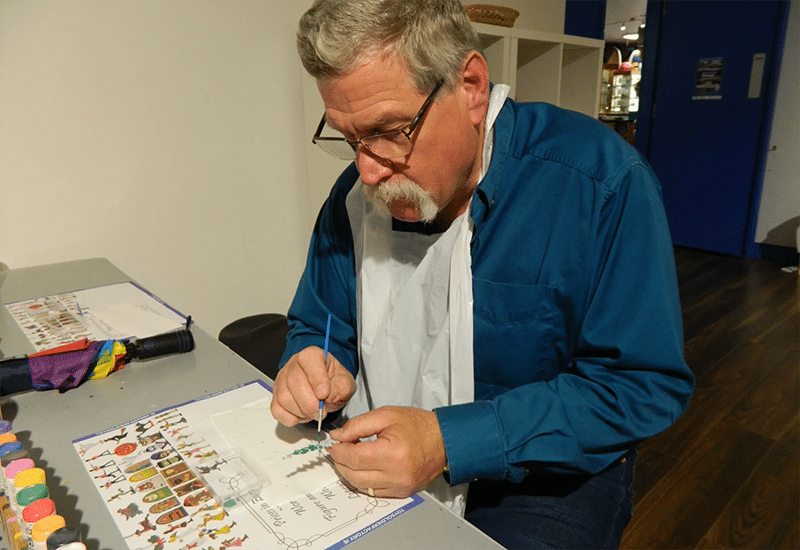 A man with gray hair and glasses, wearing a blue shirt and a white bib, is focused on painting a small piece at a table. The table is covered with various paints, brushes, and a detailed guide. The background shows a blue wall and shelves.