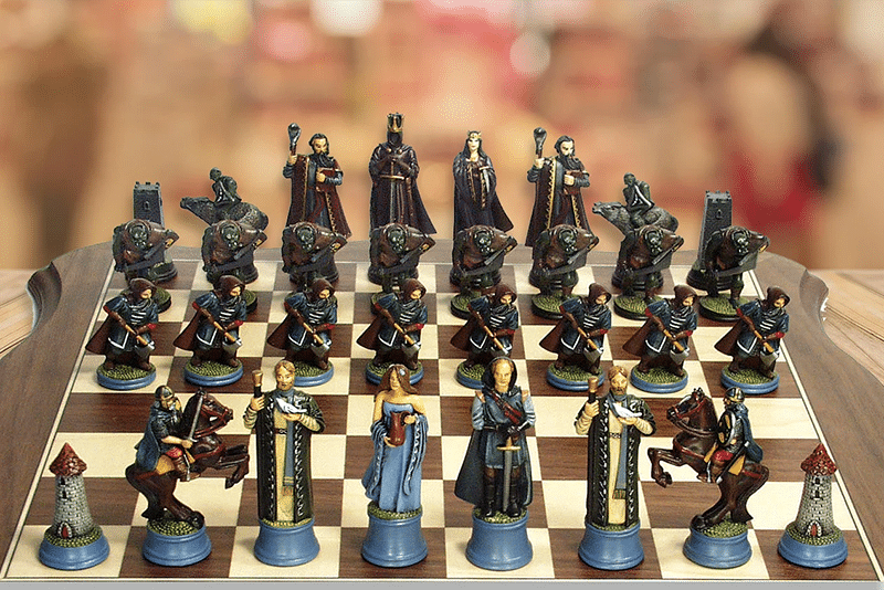 A themed chess set on a wooden board, featuring intricately detailed pieces, including knights and castles, set up and ready for a game. A blurred background suggests a public setting.