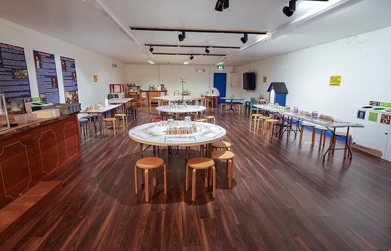 A spacious room with wooden floors and white walls, set up for educational activities. Round and rectangular tables are arranged with various crafts, models, and learning materials. Colorful chairs and stools provide seating. Information posters are on the walls.