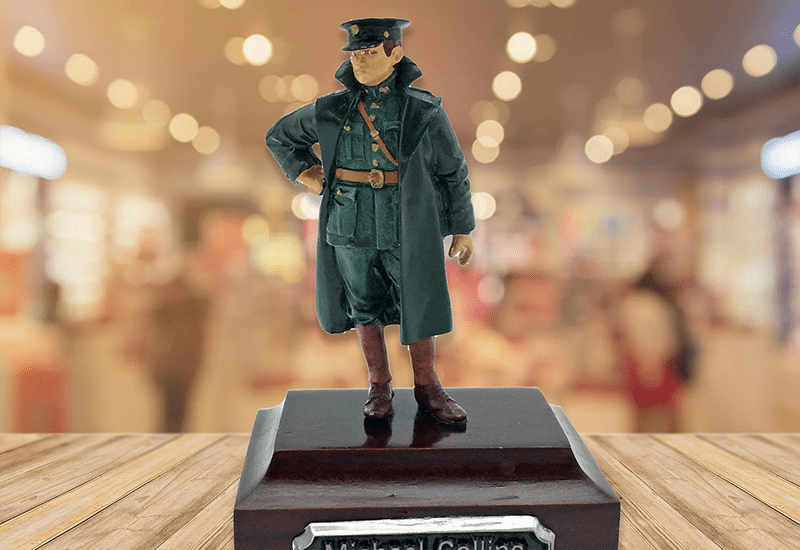 A figurine of a military officer stands on a wooden platform with a blurred indoor background. The uniform includes a cap, long coat, and knee-high boots. There is a plaque on the front of the base.