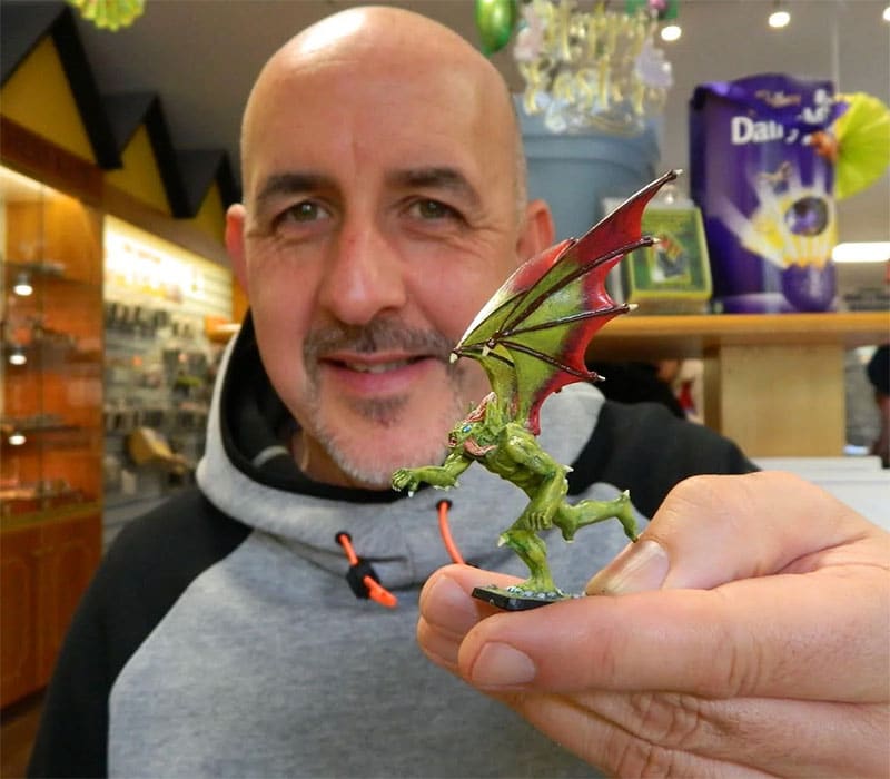 A man in a gray and black hoodie holds up a detailed miniature figurine of a green, winged creature with red-tinted wings. He is standing in a brightly lit shop, which has shelves filled with various items in the background.