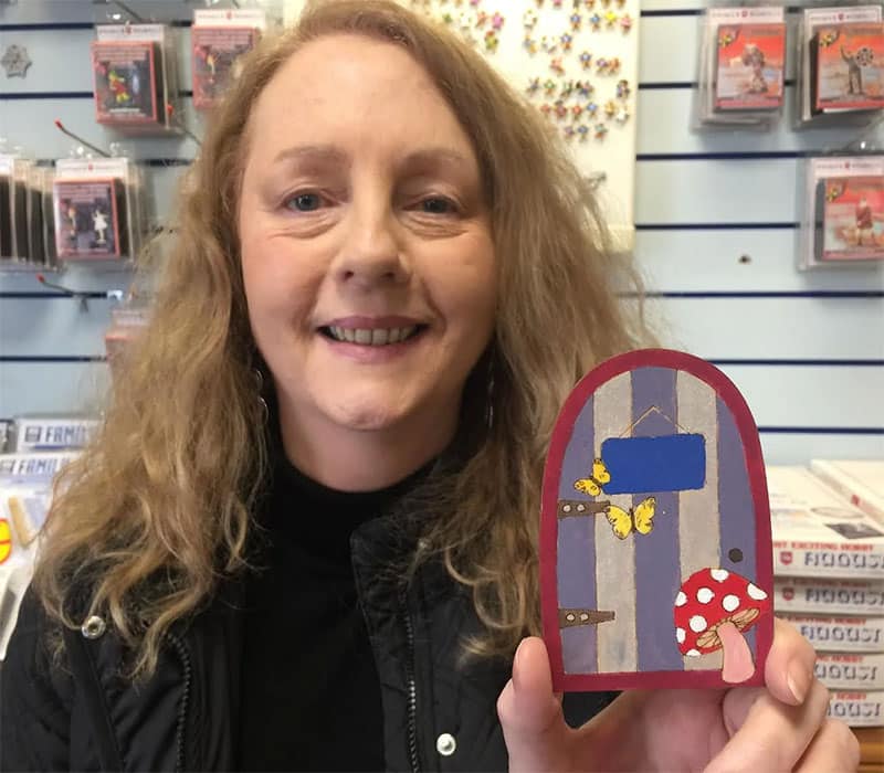 A woman with long curly blonde hair is smiling and holding a small decorative door. The door is painted with blue and purple stripes, adorned with yellow butterflies, and has a red mushroom with white spots at the base. The background displays various items on shelves.