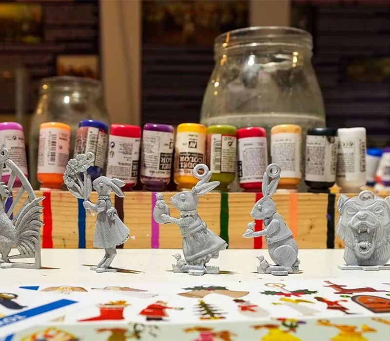 A worktable displays miniature figurines, including a rooster, two bunnies, a girl, a bear, and an elephant, all yet to be painted. Behind them are various colorful paint bottles arranged in rows, with a large glass jar in the center of the background.