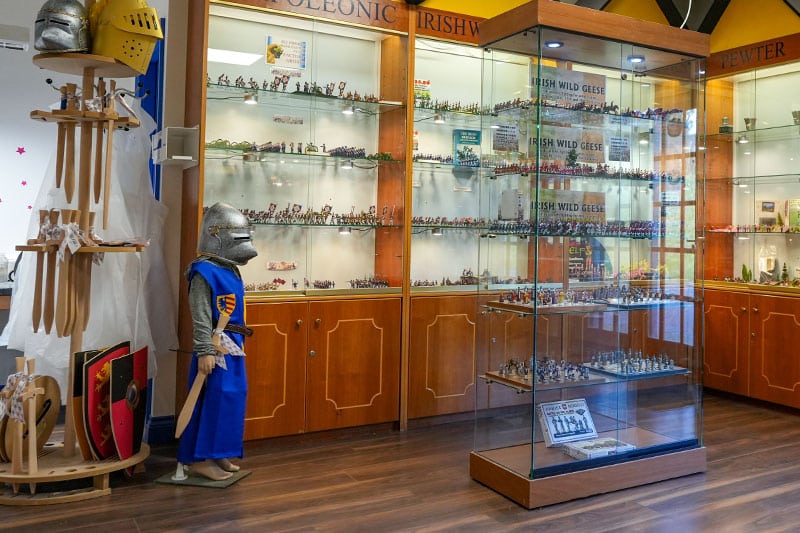 A well-lit museum exhibit displaying numerous historical figurines and miniatures in glass cabinets. To the left, there's a suit of medieval knight armor next to a rack of shields and wooden swords for children. The exhibit is titled "IRISH WILD GEESE.