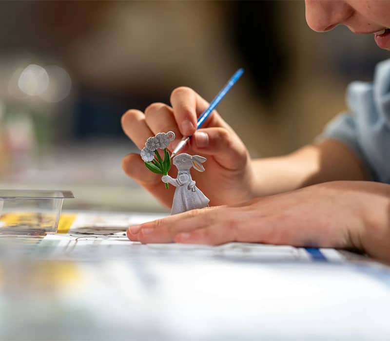 A person holds a small paintbrush, carefully painting a gray rabbit figurine that is holding a bouquet of flowers. The scene is bright and focused on the detailed work being done on the miniature figure.