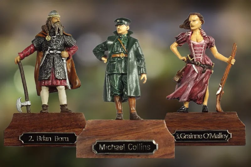 Three small figurines on wooden bases are displayed. The first portrays a medieval warrior with an axe, labeled "Brian Boru". The middle one is a figure in green military attire labeled "Michael Collins". The third is a woman with a dress and gun, labeled "Grainne O'Malley".