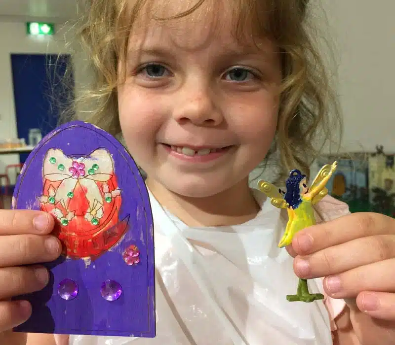 Young girl holding a painted and bejewelled wooden door with a Christmas theme, and a metal fairy.