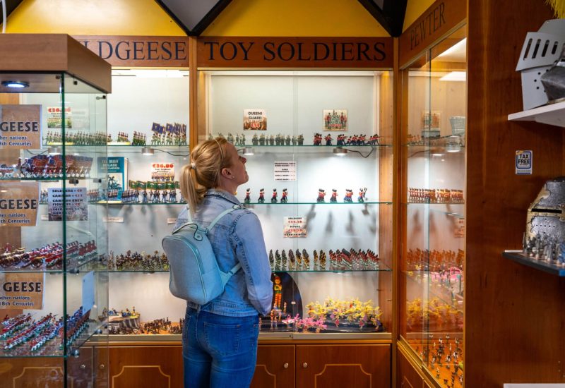 A woman with a backpack looking at various miniature toy soldiers displayed inside a glass case in a store. The store's signage reads "DODGESE TOY SOLDIERS.