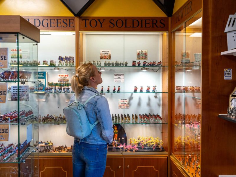 A woman wearing a backpack examines miniature toy soldiers displayed in glass cases at a store. The cases are illuminated and labeled with various descriptions.