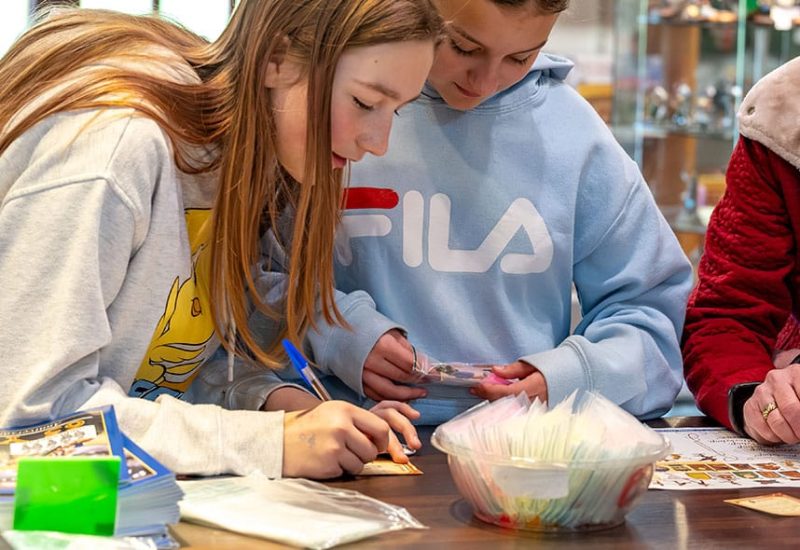 Two children are engaged in an activity at a table. One, wearing a gray hoodie, writes on a piece of paper, while the other, in a blue Fila hoodie, looks on intently. Various materials, including magazines and packets, are spread out on the table in front of them.