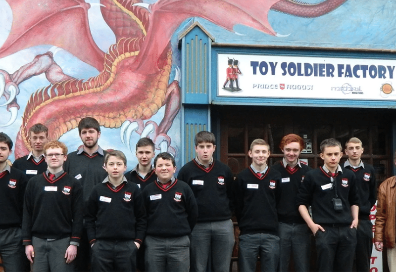 A group of young men in school uniforms stands in front of a building labeled "Toy Soldier Factory." The building features a colorful mural of a dragon. An older man stands to the far right of the group.