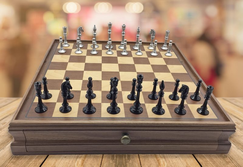 A wooden chessboard with black and silver pieces set up for a game, positioned on a table with a blurred market or mall background.